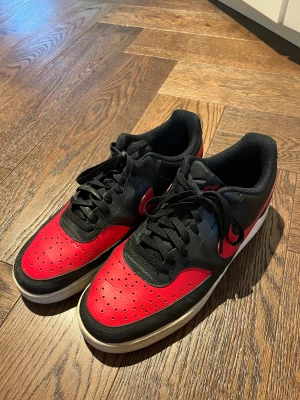 Nike Air Force 1 svart/röd sneakers - Säljer ett par Nike Air Force 1 sneakers i svart och rött med klassisk låg siluett. Skorna har perforerad tå, snörning och en vit platt sula. Swoosh-loggan syns tydligt på sidan och ovandelen är i skinn. Storlek 42