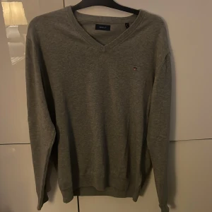 Grå v-ringad tröja från GANT XL - Grå v-ringad tröja från GANT i storlek XL. Klassisk design med ribbade muddar och diskret logga broderad på bröstet. Perfekt för dig som gillar stilrena och enkla plagg. Materialet är mjukt och känns skönt mot huden.
