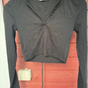 Svart croppad topp från Zara - Säljer en svart croppad topp från Zara med lång ärm och ribbad struktur. Toppen har v-ringning med knappar framtill som ger en chill och snygg look. Perfekt att matcha med jeans eller kjol för en trendig stil.