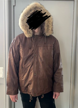 Brun skinnjacka med pälsluva från SAKI - Säljer en brun skinnjacka från SAKI med stor fluffig pälsluva och quiltat, glansigt foder. Jackan har dragkedja och knappar framtill, snörning nedtill och två snedställda fickor. Insidan är fodrad med polyester och har snygga detaljer. Perfekt för kalla dagar.