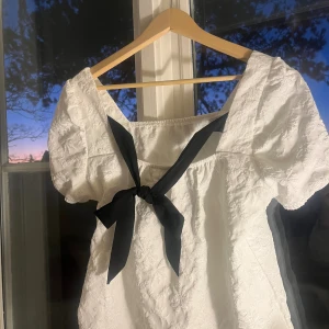 Vit blus med puffärm och svart rosett - Säljer en söt vit blus med puffärmar och broderade blommor i tyget. Blusen har en fyrkantig urringning och en svart rosett framtill som ger en extra touch. Perfekt för dig som gillar romantisk och trendig stil.