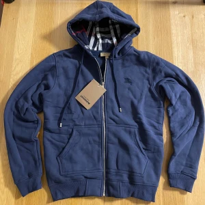 Marinblå hoodie från Burberry - Snygg marinblå hoodie från Burberry med klassiskt rutigt foder i huvan och broderad logga på bröstet. Tröjan har dragkedja, snörning i huvan och två fickor framtill. Tillverkad i mjuk bomull för skön komfort och stil.