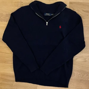 Marinblå quarter zip från Polo Ralph Lauren - Snygg marinblå stickad tröja från Polo Ralph Lauren med röd logga på bröstet. Tröjan har lång ärm, ribbade muddar och en half zip-dragkedja vid halsen. Storlek M men passar också väldigt bra som storlek S