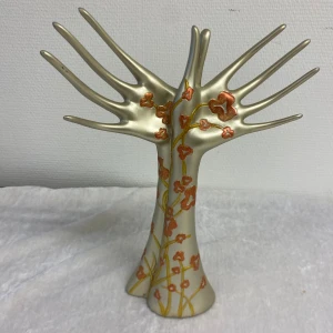 Smyckeshållare i form av händer med blommor - Unik smyckeshållare i form av två stiliserade händer i matt guld med handmålade orange blommor och slingrande gulddetaljer. Perfekt för att hänga ringar, armband eller halsband. Skulptural och dekorativ design som sticker ut på sminkbordet.