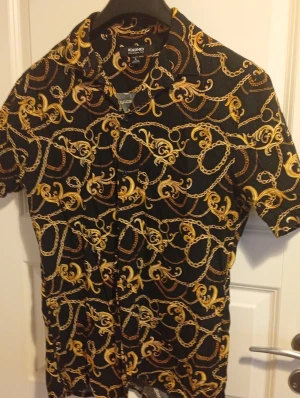 Svart skjorta med kedjemönster från Icono - Svart kortärmad skjorta från Icono i relaxed fit, täckt med ett detaljerat mönster av guldfärgade kedjor och ornament. Skjortan har klassisk krage och knappar framtill, perfekt för dig som gillar att sticka ut med coola prints.