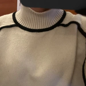 Beige polotröja med svarta detaljer - Snygg beige polotröja med svarta kontrastdetaljer längs krage, axlar, ärmslut och nederkant. Tröjan är stickad och har en ribbad hög krage samt långa ärmar. Perfekt för dig som gillar enkel men stilren design med lite edge.