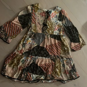 Patchwork blommig kortklänning med snörning - Superfin lpngklänning med blommiga och randiga partier samt snörning framtill. Långa ärmar och volang nedtill ger en bohemisk vibe. Perfekt för dig som gillar unika plagg med mycket färg och mönster. Storlek 134 (9år)