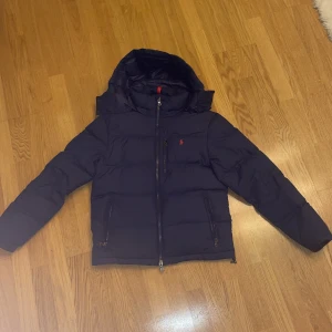 Mörkblå dunjacka från Polo Ralph Lauren - Snygg mörkblå dunjacka från Polo Ralph Lauren med huva och röd logga på bröstet. Jackan har två sidofickor med dragkedja och en central dragkedja framtill. Perfekt för kalla dagar och har en klassisk, puffad look. Perfekt julklapp till sonen eller pojkvännen 