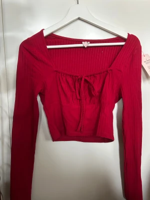 Röd croppad långärmad topp med knyt - Säljer en snygg röd croppad topp med långa ärmar och ribbad struktur. Toppen har en söt knytning framtill och en fyrkantig urringning som ger en trendig vibe. Perfekt för dig som gillar att sticka ut med färg och detaljer. Oanvänd