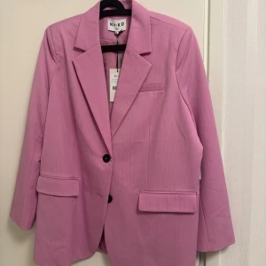 Oversized rosa kavaj från NA-KD - Snygg oversized kavaj från NA-KD i en härlig rosa färg. Klassisk design med två knappar framtill, breda slag och fickor med lock. Perfekt för dig som gillar statement-plagg och vill sticka ut med färg. Materialet är mjukt och har en lätt struktur.
