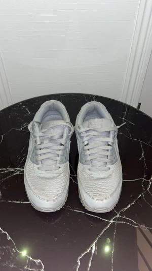 Nike Air Max 90 grå/vita sneakers - Fräscha Nike Air Max 90 sneakers i gråvitt med mesh och skinnpaneler. Klassisk chunky sula med synlig Air-enhet och snygga detaljer. Rund tå, snörning och platt sula gör dem både stilrena och bekväma. Perfekta för dig som gillar att kombinera det ihop med jeans, mjukis eller något överlag. 