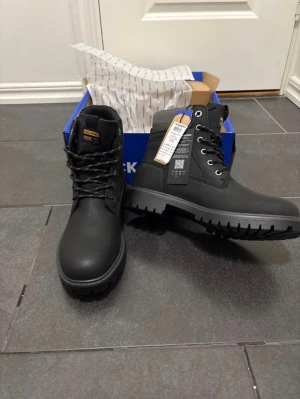 Svarta boots från Jack & Jones - Säljer ett par svarta boots från Jack & Jones med grov sula och snörning. Skorna har rund tå, grå detaljer vid skaftet och är tillverkade i syntetmaterial med nubuck-look. Perfekt för dig som gillar robust stil och vill ha något som funkar hela hösten och vintern.