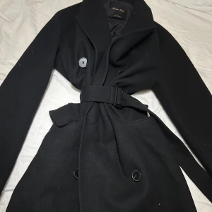 Svart kappa från Massimo Dutti - Stilren svart kappa från Massimo Dutti med klassisk krage, knappar framtill och ett snyggt bälte i midjan. Jackan har långa ärmar och är tillverkad i ullblandning som ger en lyxig känsla. Perfekt för dig som gillar minimalistisk och tidlös design.