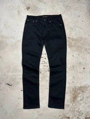 Svarta jeans från Nudie Jeans - Snygga svarta jeans från Nudie Jeans med klassisk femficksdesign och raka ben. Jeansen har svarta sömmar och Nudie-logga på bakfickan. Materialet är slitstarkt denim i bomull och passformen är normal med midwaist.
