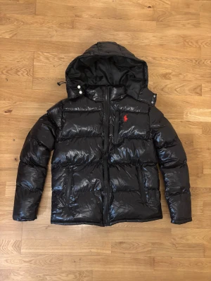 Svart pufferjacka från Polo Ralph Lauren - Säljer en svart pufferjacka från Polo Ralph Lauren med blank finish och röd broderad logga på bröstet. Jackan har huva, flera dragkedjor och snygga detaljer som snörning vid huvan. Perfekt för kalla dagar och riktigt stilren.
