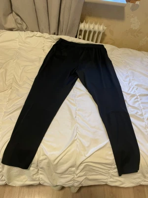 Svarta träningsbyxor från Nike - Svarta mjukisbyxor från Nike med klassisk vit logga på vänster lår. Byxorna har elastisk midja med snörning och är tillverkade i ett mjukt syntetmaterial som passar perfekt för träning. Passformen är normal och benen är raka.