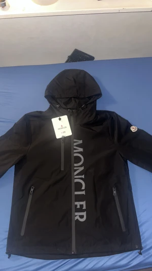 Svart vindjacka från Moncler med huva - Svart vindjacka från Moncler med stor logga längs dragkedjan, justerbar huva och två snedställda fickor med dragkedja. Jackan har klassisk Moncler-patch på ärmen och diskreta detaljer i rött, vitt och blått. Perfekt för dig som vill ha en stilren och sportig look.