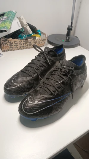 Nike Zoom Mercurial Vapor 15 Pro FG fotbollskor - Säljer ett par Nike Air Zoom Mercurial fotbollsskor i svart med blå och vita detaljer. Skorna har snörning, rund tå och platt sula med dobbar för gräsplan. Ovandelen är tillverkad i syntetmaterial med stickad känsla för extra komfort och passform. Perfekt för dig som vill ha grym touch och snabbhet på planen. Ordinarie pris 2000 kr.