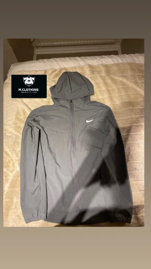 Grå Nike repel Miller windbreaker  - Snygg grå vindjacka från Nike med klassisk logga på bröstet. Jackan har hel dragkedja framtill, huva och elastiska muddar vid ärmsluten. Perfekt för blåsiga dagar och enkel att matcha med streetwear.