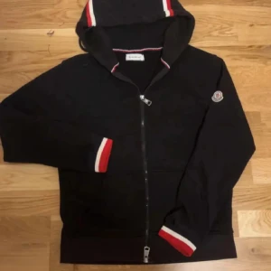 Svart Moncler hoodie med dragkedja - Svart hoodie från Moncler med hel dragkedja, huva och klassisk logga på ärmen. Rödvita detaljer på muddar och huva ger en sportig vibe. Tillverkad i mjuk bomull, perfekt för chill dagar. Fickor framtill och snygg passform.