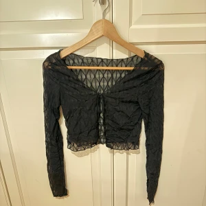 Svart mesh croppad blus med snörning - Säljer en svart mesh blus med lång ärm och croppad passform. Toppen har ett transparent rutmönster och snygg snörning framtill vid v-ringningen. 