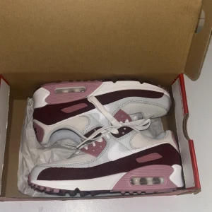 Nike Air Max 90 SE vinröd/rosa  - Nike Air Max 90 SE sneakers i vinrött, rosa och vitt. Snygg retrodesign med synlig Air Max-dämpning i sulan, snörning och platt sula. Ovandel i mesh och skinn med detaljer i mocka. Perfekt för dig som gillar streetstyle och vill sticka ut med färg. Helt oanvända 