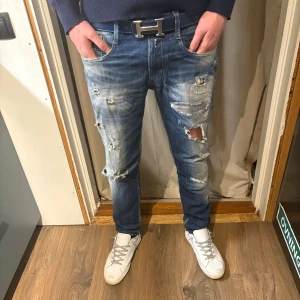Blå slitna jeans från Replay, 32/32 - Säljer ett par blå jeans från Replay med coola slitningar och hål på benen för en edgy look. Modellen har klassisk femficksdesign, normal passform och är tillverkad i jeansmaterial. Perfekt för dig som vill sticka ut i stilen!