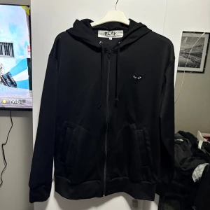 Svart hoodie från Comme des Garçons - Säljer en svart hoodie från Comme des Garçons med dragkedja och huva. Den har ett ikoniskt hjärta med ögon på bröstet och en större version på ryggen. Perfekt för en stilren look. Kom med bud!