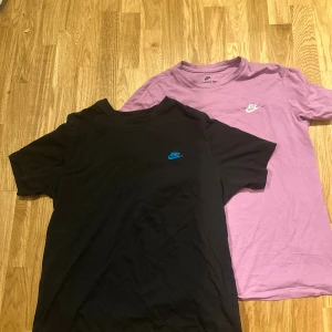 Två Nike t-shirts svart & lila. L och M - Säljer två snygga t-shirts från Nike, en i svart storlek L med blått Nike-logga och en i lila storlek M med vit logga. Båda har klassisk rund hals och korta ärmar. Perfekt för dig som gillar sportiga och stilrena plagg med ikonisk branding.
