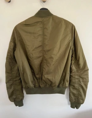 Olivgrön bomberjacka - Snygg olivgrön bomberjacka med klassisk ribbad krage, muddar och midja. Jackan har silvriga dragkedjor framtill och på ärmen samt två snedställda fickor.