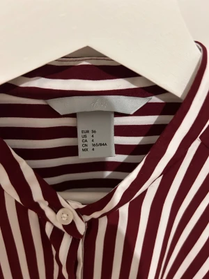 Randig skjorta från H&M, strl 36 - Snygg skjorta från H&M i storlek 36 med vertikala vinröda och vita ränder. Skjortan har klassisk krage, långa ärmar och knäppning framtill. Tillverkad i mjukt och lätt polyestermaterial som känns skönt mot huden. Perfekt för dig som gillar stilrena mönster och färger.