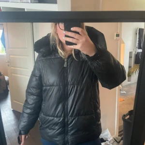 Svart pufferjacka från Guess, M - Säljer en svart pufferjacka från Guess i storlek M. Jackan har en stor huva, dragkedja framtill och två fickor med dragkedja. 