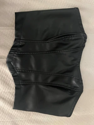 Svart kort tub topp  - Snygg svart tub top från SHEIN i storlek XS. Tub toppen är tillverkad i syntetmaterial med en skinnliknande finish och har markerade sömmar för en cool look. Dragkedja baktill gör den enkel att ta på och av. Perfekt för dig som gillar edgy stil.