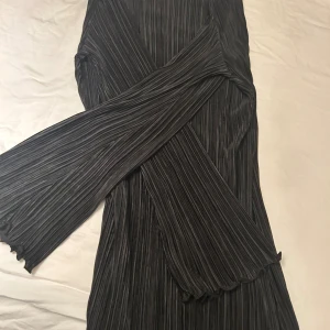 Svart långärmad plisserad klänning - Snygg svart långklänning med plisserat tyg och långärmade ärmar. Klänningen har ett randigt mönster och en smickrande omlott-design framtill. Materialet känns mjukt och har ett fint fall, perfekt för dig som gillar stilrena och moderna plagg.