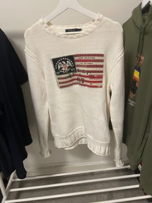 Vit stickad tröja Polo Ralph Lauren - Vit stickad tröja från Polo Ralph Lauren med amerikansk flaggpatch och text på bröstet. Tröjan är i storlek M men skulle säga att den litr lös i sin passform så att den lär passa större storlekar också. Skicket är 10/10 jag har alldrig använt den. 