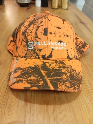 Orange camo keps Mellanskog - Säljer en orange keps från Mellanskog med coolt camouflage-mönster i brunt och svart. Kepsen har böjd skärm och broderad logga framtill. Perfekt för dig som gillar skog och outdoor-vibe.