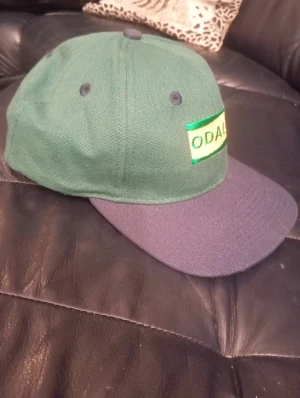Odal keps - Snygg keps från Odal med grön krona och mörkblå skärm. Framtill finns ett broderat Odal-märke i grönt och gult. Kepsen har klassisk form med ventilationshål och är tillverkad i bomull. Perfekt för dig som vill ha en chill och stilren look.