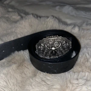Cowboy Svart bälte med silverfärgat oxspänne - Säljer ett svart bälte i skinn med detaljerat präglat mönster längs hela remmen. Spännet är stort, silverfärgat och har en cool oxdesign med ornamenterade detaljer. Perfekt för dig som vill ha western-vibe och sticka ut med accessoarer.