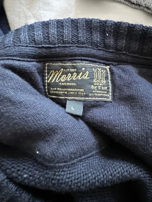 Mörkblå stickad tröja Morris L - Snygg mörkblå stickad tröja från Morris i storlek L. Tröjan har ett klassiskt kabelstickat mönster framtill och ribbade muddar vid ärmslut och nederkant. På bröstet finns en broderad patch med brittisk flagga och text. Perfekt för dig som gillar stilren och tidlös design.