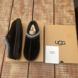 Svarta UGG slip-ons med mönstrad kant - UGG slip-ons i svart mocka med rund tå och platt sula. Skorna har en vit zigzag-broderad kant runt öppningen och mjukt, ljust foder inuti för extra komfort. Perfekt för dig som gillar stilren design med en twist.