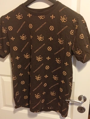 Svart Icono Couture t-shirt M - Svart t-shirt från Icono Couture i storlek M med bruna tryck över hela plagget. T-shirten har korta ärmar och rund hals. Mönstret består av loggor, text och dekorativa symboler. Materialet är mjuk bomull och passformen är normal.