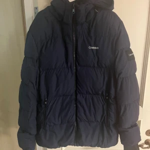 Mörkblå pufferjacka från Calvin Klein - Snygg mörkblå pufferjacka från Calvin Klein med huva och dragkedja framtill. Jackan har två fickor med dragkedja och logga på bröstet samt ärmen. Perfekt för kalla dagar och riktigt stilren look. Storlek XL priset går o diskutera 