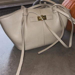 Beige handväska med gulddetalj - Snygg beige handväska i skinn med stilren form och långa remmar. Väskan har en cool guldfärgad vridlåsdetalj framtill och minimalistisk design. Perfekt storlek för att få plats med allt du behöver och passar till många olika looks.