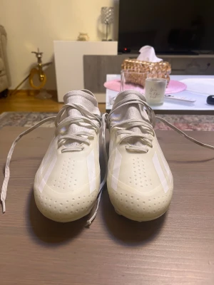 Adidas X Crazyfast vita fotbollsskor - Säljer ett par Adidas X Crazyfast fotbollsskor i helvitt med klassiska tre ränder på sidorna. Skorna har snörning, platt sula med dobbar och en strumpliknande ovandel för extra komfort. Perfekta för dig som vill sticka ut på planen med stilrena och snabba skor.