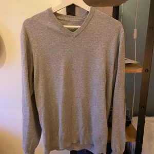 John Henric Grå v-ringad stickad tröja - En enkel och stilren grå stickad tröja med v-ringad hals. Tröjan har långa ärmar och ribbade muddar vid hals, ärmslut och nederkant. Perfekt för dig som gillar en clean look och vill ha något mjukt och bekvämt.