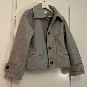 Grå kappa  - Snygg grå overshirt från KappAhl med klassisk krage, stora framfickor och knappar framtill. Jackan har lång ärm med knappdetalj vid mudden och är tillverkad i mjuk ullmix. Perfekt för lager-på-lager under höst och vår.