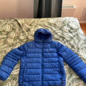 Blå pufferjacka från Nicce med huva - Säljer en blå pufferjacka från Nicce med huva och dragkedja framtill. Jackan har quiltad design och två sidofickor med dragkedja. Perfekt för kalla dagar och har en clean look med diskret logga på bröstet. Hör av dig vid minsta lilla fråga:)