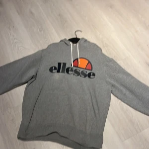 Grå Ellesse hoodie XL med logga - Säljer en grå hoodie från Ellesse i storlek XL. Tröjan har stor tryckt logga framtill i svart, orange och rött, samt vit snörning i huvan. Klassisk passform med långa ärmar och mjukt material, perfekt för chill dagar.