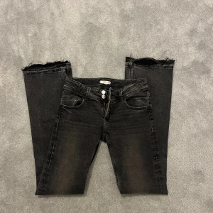 Svarta bootcut jeans från Ginatricot - Snygga svarta jeans från Ginatricot i bootcut-modell med låg midja. Jeansen har slitna, råa kanter vid bensluten och två knappar i midjan. Materialet är klassisk jeans med lite stretch för skön passform.