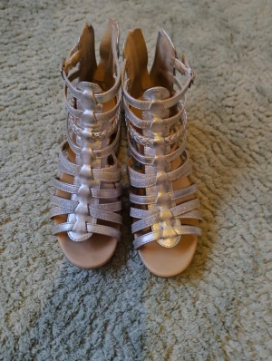 Silvriga sandaler från Zoey, strl 37 - Säljer ett par glittriga silvriga sandaler från Zoey i storlek 37. De har flera remmar, flätade detaljer och öppen tå. Dragkedja baktill gör dem enkla att ta på.